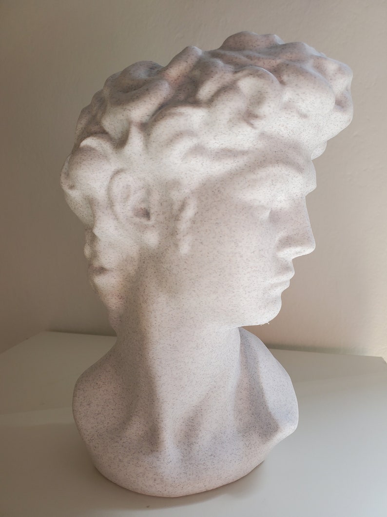 May include: A white plaster bust sculpture of a person with a textured surface. The bust is of a person's head and shoulders, with the person's hair styled in a short, wavy style.