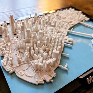 Op de afbeelding: Een wit 3D-geprint model van de skyline van Manhattan, in een zwart frame met een blauwe achtergrond. Het model bevat de iconische wolkenkrabbers en bruggen van de stad. Het frame heeft de tekst "Manhattan" gedrukt in de rechterbenedenhoek.