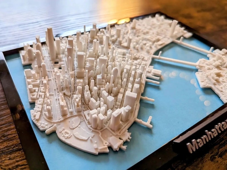 Op de afbeelding: Een wit 3D-geprint model van de skyline van Manhattan, in een zwart frame met een blauwe achtergrond. Het model bevat de iconische wolkenkrabbers en bruggen van de stad. Het frame heeft de tekst "Manhattan" gedrukt in de rechterbenedenhoek.