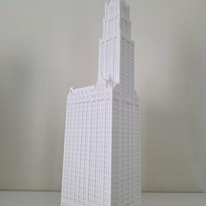 May include: A white architectural model of a skyscraper with a detailed facade. The building has a tall, central tower and a stepped design, with numerous windows and architectural details. The model is set against a plain white background.
