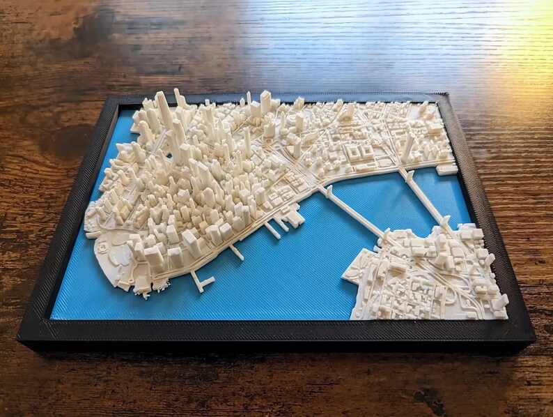 Op de afbeelding: Een 3D-geprint model van een stadsbeeld, met witte gebouwen en een blauwe waterbasis. Het model is ingelijst in zwart en wordt weergegeven op een houten oppervlak.