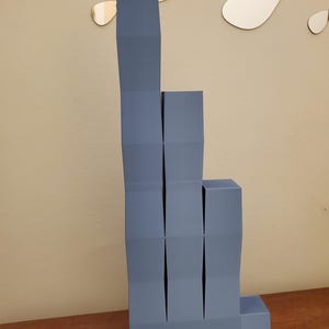 May include: A modern, abstract sculpture in shades of blue-gray. The geometric design features stacked, folded rectangular forms, creating a stepped, architectural appearance. The sculpture is placed on a wooden surface.