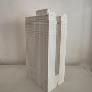 Peut inclure: Une maquette architecturale blanche imprimée en 3D d'un gratte-ciel présentant une base rectangulaire, une section centrale en retrait et plusieurs niveaux de motifs de fenêtres répétitifs sur la façade, surmontée d'un toit plat.