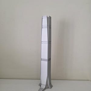 Può includere: Modello architettonico color argento di un grattacielo. Il modello ha una forma rettangolare con un design affusolato e una facciata dettagliata. La base del modello è più larga della parte superiore e si erge su una superficie piana.