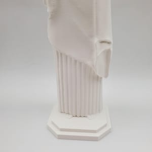 May include: White plaster statue of a woman standing on a square base with a fluted column. The statue is missing a portion of the drapery.