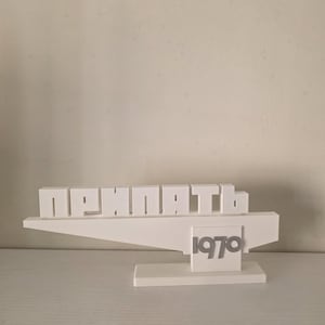 May include: A white, 3D-printed sculpture featuring the word "ПРИПЯТЬ" in block letters and the number "1970". The sculpture has a geometric design with a horizontal plane and a vertical base. The background is a plain, off-white wall.