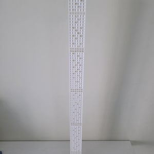 May include: A white architectural model of a skyscraper. The model is tall and rectangular, with numerous small, square windows. The building is constructed from a light-coloured material and stands upright. The background is a plain, light grey.