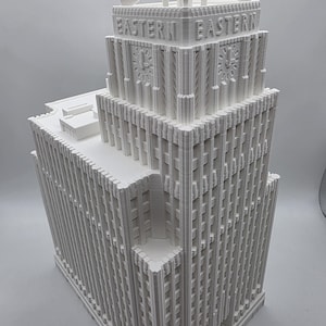 May include: A white, 3D-printed model of a skyscraper with the word "EASTERN" on the top. The building has a detailed facade with vertical lines and clock faces. The model is a detailed architectural replica.
