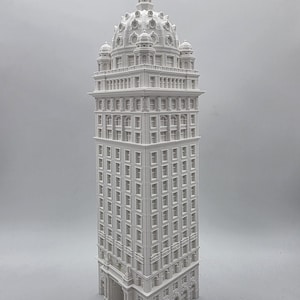 May include: A detailed, white architectural model of a tall building with a domed top. The model features numerous windows and intricate architectural details, showcasing a classic skyscraper design. The building's facade is a uniform white color.