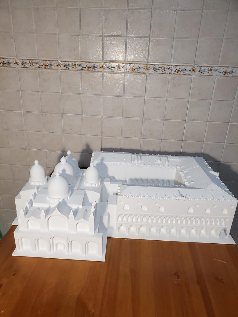 May include: A white 3D printed model of a building with multiple domes and arches. The model is on a wooden surface.