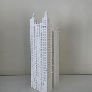 May include: A white architectural model of a tall building with many windows and two domed towers. The model is a detailed replica of a skyscraper, showcasing its intricate design and structure. The building is set against a plain background.