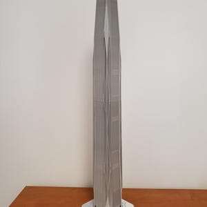 Pode incluir: Modelo de arranha-céus prateado com uma fachada detalhada e um topo pontiagudo. O modelo é montado numa base quadrada branca e numa superfície de madeira.