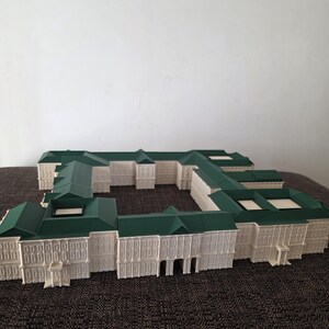 May include: A detailed architectural model of a building with a white facade and green roof. The model features multiple sections, with intricate window and door details. The building is set on a dark brown surface.