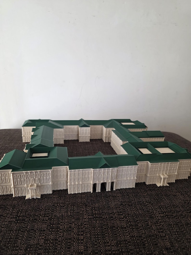 May include: A detailed architectural model of a building with a white facade and green roof. The model features multiple sections, with intricate window and door details. The building is set on a dark brown surface.