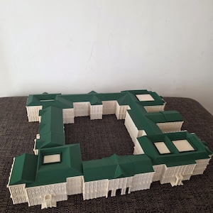 May include: A white and green architectural model of a building complex. The model has a green roof and white walls with detailed windows and architectural elements. The model is placed on a brown surface.