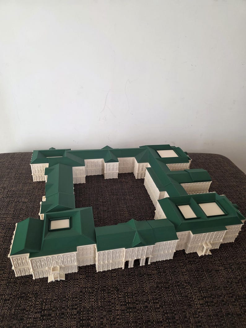 May include: A white and green architectural model of a building complex. The model has a green roof and white walls with detailed windows and architectural elements. The model is placed on a brown surface.