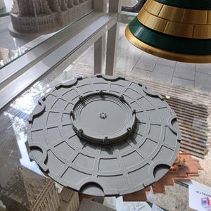 Può includere: Un oggetto circolare grigio stampato in 3D caratterizzato da un disco centrale rialzato circondato da un motivo a griglia con bordi dentellati. La superficie presenta una texture a strati tipica della produzione additiva, con piccoli dettagli simili a bulloni.