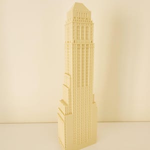 Può includere: Modello beige di un grattacielo. L'edificio presenta una facciata dettagliata con numerose finestre e un design a gradini. Il modello è di colore beige chiaro e si erge su uno sfondo neutro. Replica in miniatura.