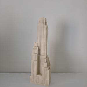 Könnte beinhalten: Beiges Architekturmodell eines Wolkenkratzers. Das Gebäude hat ein gestuftes Design mit mehreren rechteckigen Abschnitten und einer detaillierten Fassade. Das Modell steht auf einer weißen Oberfläche vor einem schlichten weißen Hintergrund.