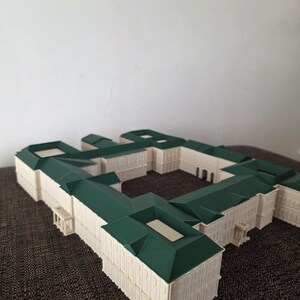 May include: A detailed architectural model of a building, featuring a cream-coloured facade and green roofs. The model has a complex design with multiple wings and a central courtyard. The structure showcases intricate details and a symmetrical layout.