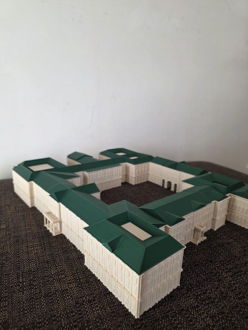 May include: A detailed architectural model of a building, featuring a cream-coloured facade and green roofs. The model has a complex design with multiple wings and a central courtyard. The structure showcases intricate details and a symmetrical layout.