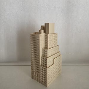 Könnte beinhalten: Ein beiges, 3D-gedrucktes Architekturmodell eines Wolkenkratzers mit abgestuftem Design. Das Bauwerk besteht aus einem hohen Hauptturm mit zahlreichen rechteckigen Fenstern und einem kleineren, niedrigeren Abschnitt an der Seite.