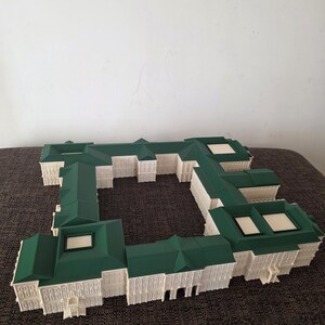 May include: A white and green architectural model of a building with a complex, rectangular design. The model features a green roof and white walls with detailed window and door features. The model is placed on a brown surface.