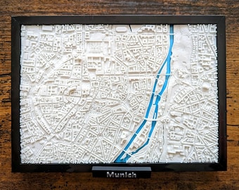 May include: A white 3D printed map of Munich, Germany, in a black frame with a blue river running through it. The text "Munich" is printed on a black plaque below the frame.