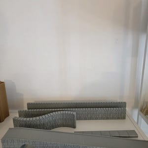 Może przedstawiać: Kolekcja szarych miniaturowych segmentów muru z kamienia oraz konstrukcja mostu. Elementy posiadają szczegółową teksturę cegieł. W zestawie znajdują się proste odcinki muru, zakrzywiony odcinek muru oraz most z gładką powierzchnią rampy.