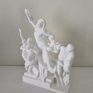 Può includere: Una scultura bianca raffigurante una scena classica con più figure. La figura centrale, un uomo barbuto, alza il braccio. Altre figure sono posizionate intorno a lui, creando una composizione dinamica. La scultura è su una base rettangolare.