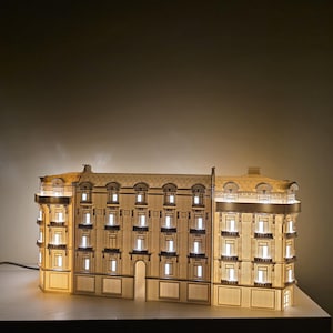 May include: A detailed, illuminated model of a building, with multiple windows and architectural features. The warm glow of the interior lights contrasts with the exterior's beige and brown tones. The model is a decorative lamp.
