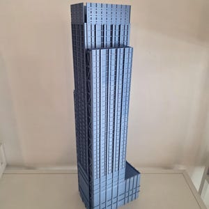 May include: A tall, light blue architectural model of a skyscraper featuring a grid-like facade with vertical and horizontal lines. The structure has a stepped design with multiple levels and a rectangular base, showcasing a detailed exterior surface.