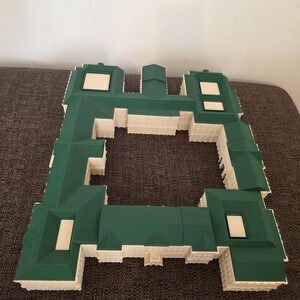 May include: A green and white architectural model of a building. The model has a complex roof design with a green roof and white accents. Rectangular windows are visible. The structure is set against a neutral background.