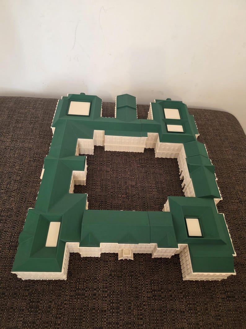 May include: A green and white architectural model of a building. The model has a complex roof design with a green roof and white accents. Rectangular windows are visible. The structure is set against a neutral background.
