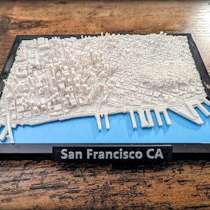 May include: A 3D relief map of San Francisco, CA, in a black frame. The map features white buildings and streets, a blue bay, and a black label with the city's name. The map is a detailed representation of the city's layout.