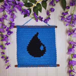 May include: A blue, crocheted wall hanging with a black teardrop design. The banner is suspended by a wooden dowel and blue yarn. Purple wisteria flowers and green leaves frame the wall hanging.