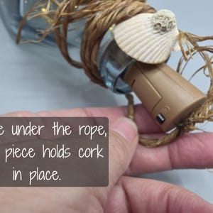 May include: A decorative bottle stopper with a seashell accent. The stopper is brown and has a switch. The stopper is wrapped with rope. The text on the image says "Wire under the rope, end piece holds cork in place."