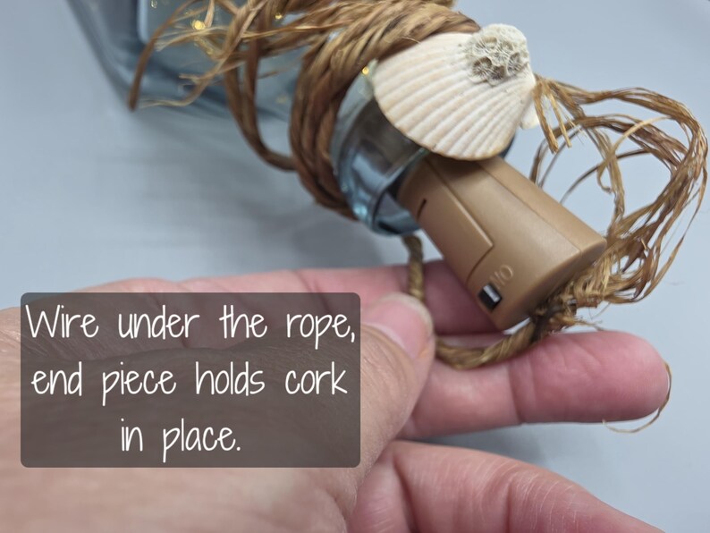 May include: A decorative bottle stopper with a seashell accent. The stopper is brown and has a switch. The stopper is wrapped with rope. The text on the image says "Wire under the rope, end piece holds cork in place."