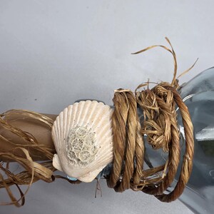 May include: A decorative bottle with a seashell and rope embellishment. The bottle is adorned with a cream-colored seashell, brown rope, and raffia. The design evokes a nautical or beach theme.