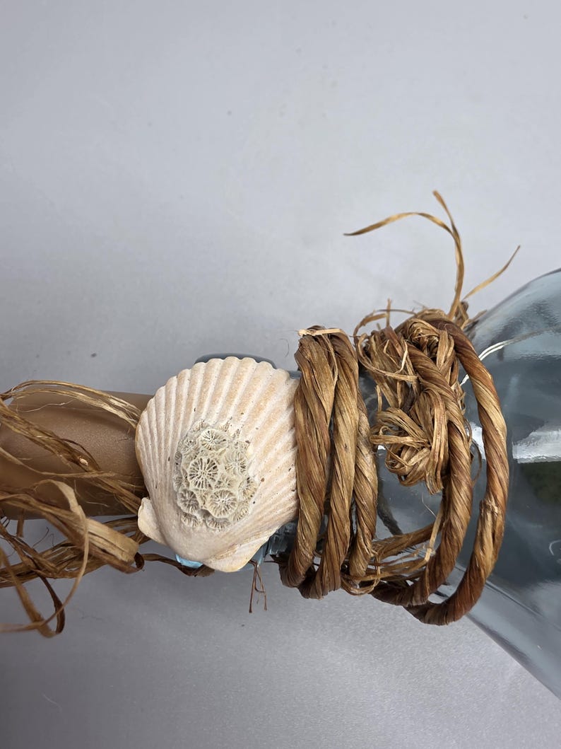 May include: A decorative bottle with a seashell and rope embellishment. The bottle is adorned with a cream-colored seashell, brown rope, and raffia. The design evokes a nautical or beach theme.