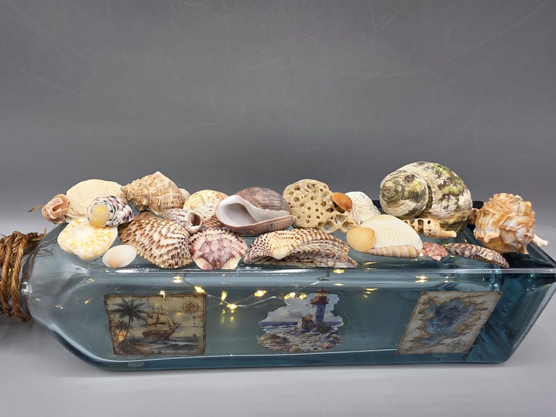 May include: A decorative glass bottle filled with seashells and fairy lights. The bottle has a blue tint and features nautical-themed images. The bottle's neck is wrapped with twine, and various seashells are arranged on top.