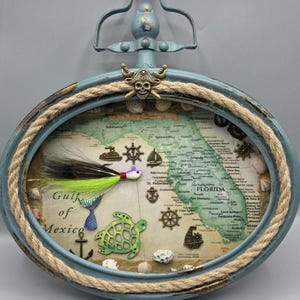 May include: An oval-shaped, light blue framed artwork featuring a map of Florida. The frame is adorned with rope and decorative elements like a pirate skull, seashells, and nautical symbols. A fishing lure and a green turtle are also included.