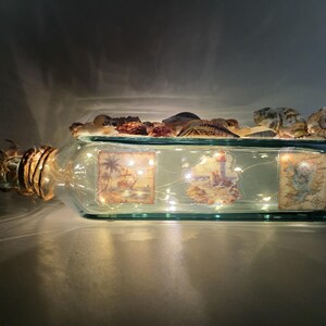 May include: A rectangular glass bottle with a cork and decorative seashells. The bottle contains fairy lights and three vintage-style nautical images. The bottle is lit up, creating a warm, inviting glow.