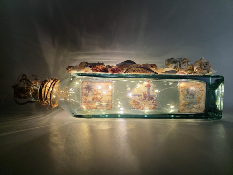 May include: A rectangular glass bottle with a cork and decorative seashells. The bottle contains fairy lights and three vintage-style nautical images. The bottle is lit up, creating a warm, inviting glow.