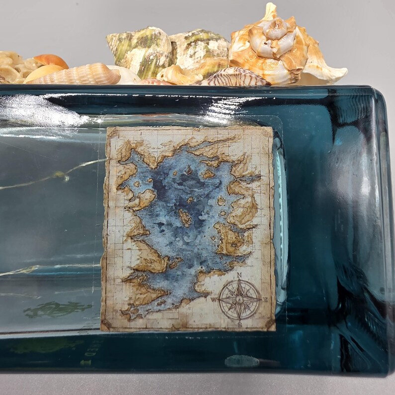 May include: A rectangular teal glass container with a vintage map design and seashells. The map features a blue body of water surrounded by tan landmasses. Seashells of various sizes and colors are arranged on top of the container.