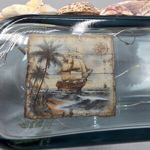 May include: A decorative glass bottle with a nautical theme. The bottle features a painted image of a sailing ship, palm trees, and a compass rose. The bottle is adorned with seashells and fairy lights.