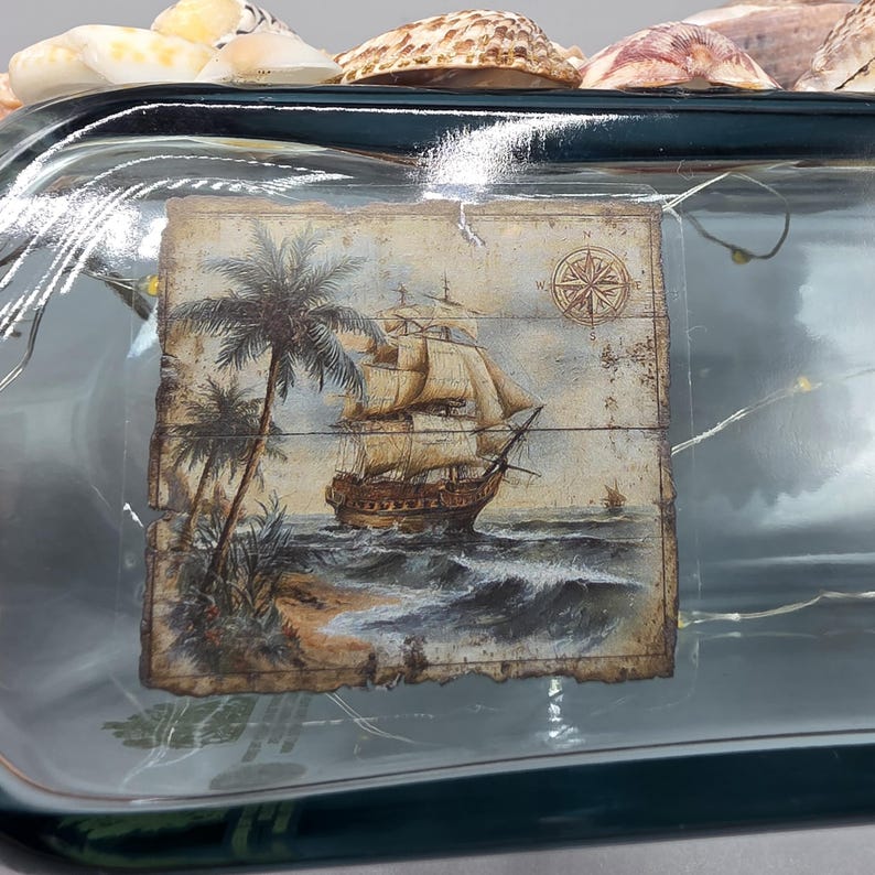 May include: A decorative glass bottle with a nautical theme. The bottle features a painted image of a sailing ship, palm trees, and a compass rose. The bottle is adorned with seashells and fairy lights.