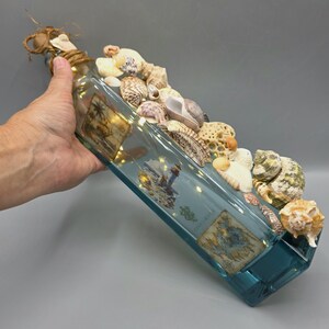 May include: A rectangular glass bottle with a blue tint, decorated with seashells and small lights. The bottle is adorned with various types of seashells and has small, warm-toned lights inside. The top is wrapped with twine.