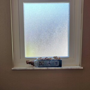 May include: A decorative blue glass bottle with a nautical theme, filled with small seashells, sits on a windowsill. The bottle has a cork stopper and is decorated with images of sailboats. The window is white and has a frosted glass pane.