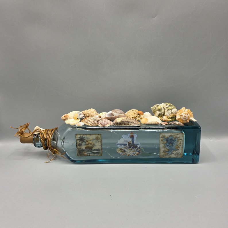 May include: A rectangular, light blue glass bottle decorated with seashells and nautical-themed images. The bottle's opening is sealed with a cork and twine. The top of the bottle is covered with various seashells.